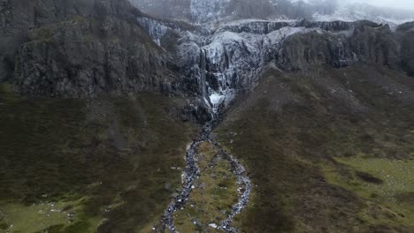 Frosty-waterfall-on-the-way-down-the-east-coast-in-the-mountains-of-Iceland,-aerial