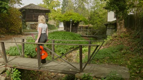 Young-female-guitarist-walks-calmly-wooden-bridge-rustic-village-scene