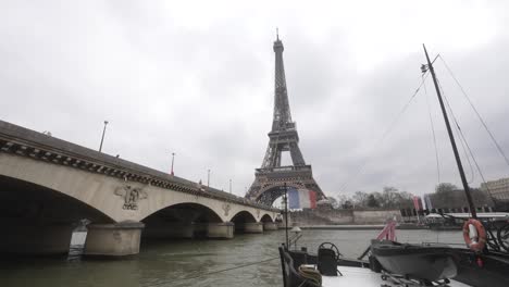 Eiffel-Tower-view-from-the-riverbanks,-a-French-flag-on-a-floating-barge,-with-the-Pont-d'Iéna-bridge-on-the-left