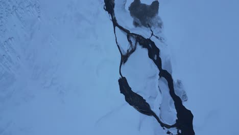 Top-down-of-a-cold-and-snowy-river-in-the-North-of-Iceland-in-the-winter,-aerial