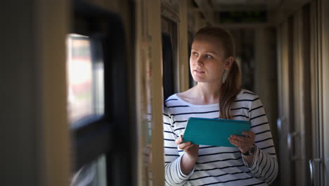 Young-woman-with-pad-standing-by-the-window-in-train-hallway