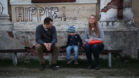 Parents-and-their-child-sitting-on-the-bench-near-old-grungy-building