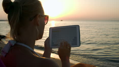Woman-typing-on-pad-sitting-by-sea-at-sunset