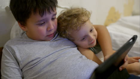 Two-boys-in-bed-with-touch-pad-One-playing-game-another-watching