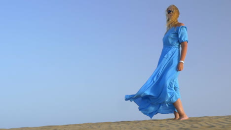 Slow-motion-view-of-young-blond-woman-standing-against-blue-sky-in-long-blue-dress-and-it-is-fluttering-on-the-wind