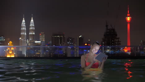 Blick-Auf-Eine-Frau-Im-Schwimmbad-Auf-Dem-Dach-Des-Wolkenkratzers-Mit-Tablet-Vor-Der-Nächtlichen-Stadtlandschaft-Von-Kuala-Lumpur,-Malaysia