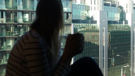 Woman-at-the-window-with-mug-looking-city-modern-glass-building
