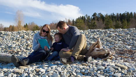 Family-of-three-sitting-on-the-stony-shore-and-watching-something-pad