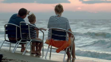 Family-of-three-sitting-on-chairs-by-sea-at-sunset