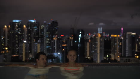 Night-view-of-young-beautiful-couple-in-swimming-pool-on-the-roof-on-foreground-and-city-landscape-Kuala-Lumpur-Malaysia