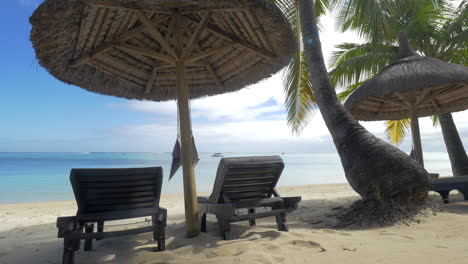 View-of-empty-chaise-longue-near-native-sun-umbrella-and-palm-trees-against-blue-water-Mauritius-Island
