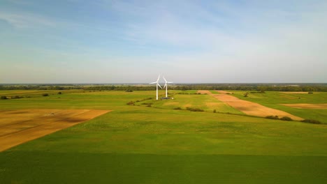 Drone-shot-of-two-working-wind-turbines-generating-green-electric-energy-on-a-cultivated-field-on-a-sunny-summer-day,-use-of-renewable-resources-of-energy,-still-shot