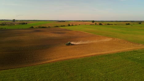 Drone-shot-of-a-green-John-Deere-tractor-preparing-the-field-for-plantation-with-a-cloud-of-dust-behind-tractor-on-a-sunny-summer-day,-following-shot