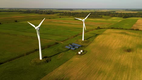 Drone-shot-of-two-working-wind-turbines-and-a-few-solar-panels-producing-green-electric-energy-on-a-cultivated-field-on-a-sunny-summer-day,-use-of-renewable-resources-of-energy,-zoom-out