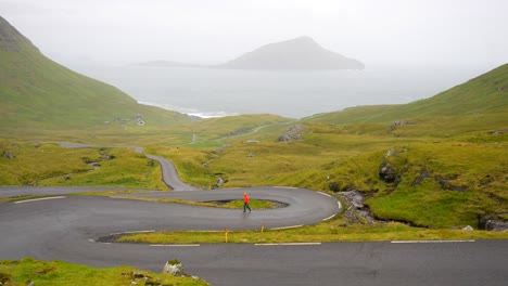 Wide-shot-of-man-walking-on-Nordradalsskard-twisty-Faroese-road-on-a-gloomy-day,-Koltur-Island-behind
