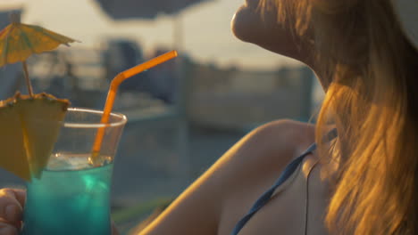 Young-woman-having-cocktail-on-beach-at-sunset
