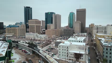 Winnipeg-Manitoba-Canada-Downtown-Skyscraper-Buildings-in-City-Overcast-Landscape-Skyline-Snowing-Winter-Drone-4k-Drone-Shot-with-Multicolored-Train-Historic-Landmark-District-and-Baseball-Arena