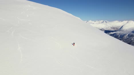 Mountaineer-walking-uphill-snowy-mountain-during-sunny-day-in-winter---Norway,Europe--Drone-shot