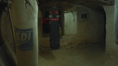 Boxer-strides-into-gritty-gym,-wearing-gloves-and-hoodie-surrounded-by-punching-bags