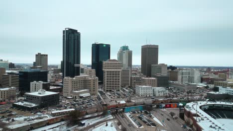 Slow-Establishing-Orbit-Winnipeg-Manitoba-Canada-Downtown-Skyscraper-Buildings-in-City-Overcast-Blue-Sky-Landscape-Skyline-Snowing-Winter-Drone-4k-Shot-with-Multicolored-Train-Passing-Through