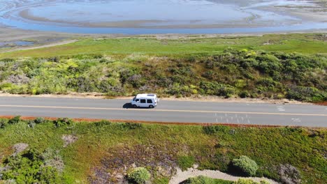 Drone-video-of-a-white-van-traveling-along-a-road-with-a-saline-in-the-backdrop,-drone-follows-the-van-before-ascending-to-reveal-the-vastness-of-the-saline,-In-Bodega-Bay,-California