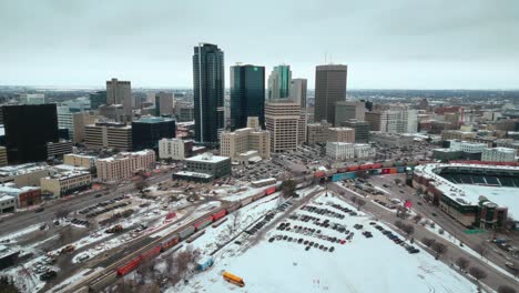 Winnipeg-Manitoba-Canada-Downtown-Skyscraper-Buildings-in-City-Overcast-Landscape-Skyline-Snowing-Winter-Drone-4k-Drone-Shot-with-Multicolored-Train-Canadian-Museum-for-Human-Rights-and-Baseball-Arena