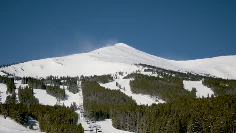 Snow-blowing-off-the-top-of-a-mountain-at-a-ski-resort-on-a-cloudless-day,-static