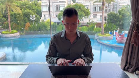 Focused-Asian-Millennial-Man-Nodding-to-Music-While-Working-on-Laptop-in-Luxurious-Home-Overlooking-Pool