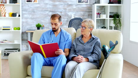 Male-assistant-in-nursing-home-reading-a-book-to-retired-elderly-pensioner