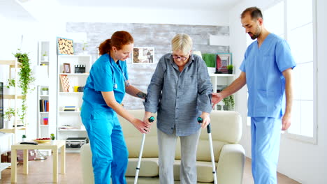 Female-doctor-and-her-assistant-helping-old-woman-with-crutches-to-stand-up-from-the-couch