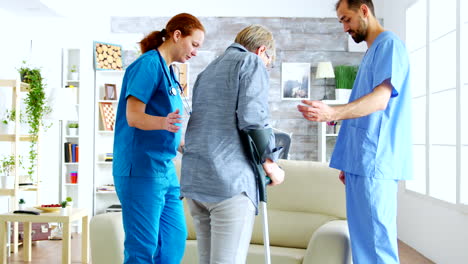 Male-doctor-and-his-assistant-in-nursing-home-helping-senior-woman-who-walk-in-in-crutches