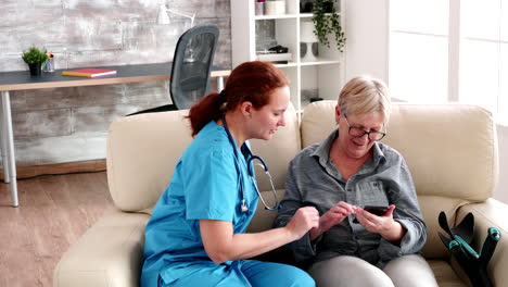 Nurse-in-retirement-home-sitting-on-the-couch-helping-elderly-woman-to-use-a-smartphone