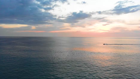 Strafing-aerial-drone-shot-of-a-beautiful-sunset-across-the-Andaman-Sea-horizon,-with-a-jetski-passing-by-located-at-the-tip-of-Phuket-in-Southern-Thailand