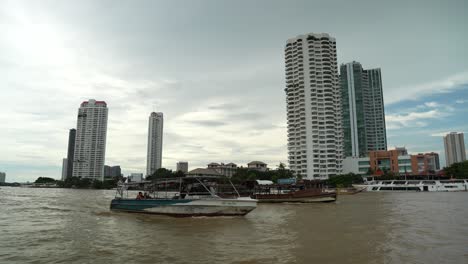 Rudimentäre-Boote-Segeln-Mit-Der-Skyline-Von-Bangkok-Am-Flussufer-An-Einem-Bewölkten-Tag