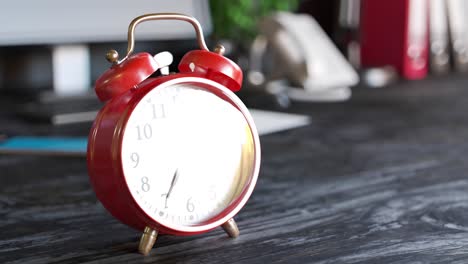 Old-vintage-red-alarm-clock-standing-on-a-wooden-office-desk.-Bells-ring-and-clock-shakes-on-time.-Office-workstation-in-a-background.-Symbol-of-morning,-countdown,-deadline.--Classic-ageless-design.