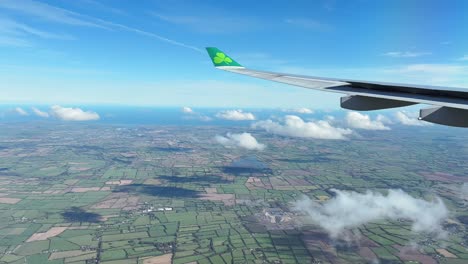 Aer-Lingus-plane-wing-in-view-while-plane-is-landing-over-green-fields,-blue-ocean-and-puff-clouds-in-Dublin,-Ireland
