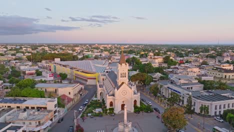 Aerial-orbit-shot-of-CATHEDRAL-SAN-PEDRO-APOSTOL-during-golden-hour-in-SAN-PEDRO-DE-MACORIS,-Dominican-Republic---People-on-square-with-monument