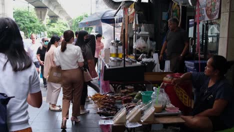 A-vendor-fanning-herself-from-the-heat-produced-by-the-morning-sun-on-the-street-as-she-sells-snacks-and-barbecued-food-to-people-passing,-Street-Food-along-Sukhumvit-Road-in-Bangkok,-Thailand