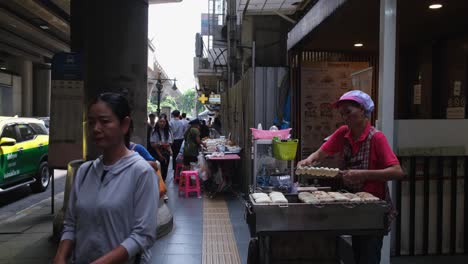 Frau-Mit-Rotem-Hemd-Verkauft-Zusammengebratene-Bananen-Auf-Einem-Bürgersteig-Und-Menschen-Kommen-Und-Gehen,-Im-Hintergrund-Ist-Auch-Eine-Apotheke-Zu-Sehen,-Streetfood-Entlang-Der-Sukhumvit-Road-In-Bangkok,-Thailand
