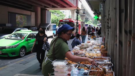 Vendor-stirring-and-arranging-food-in-containers-preparing-for-lunchtime-along-Sukhumvit-road-as-traffic-progresses,-Street-Food-along-Sukhumvit-Road-in-Bangkok,-Thailand