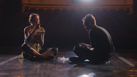Pair-of-weary-classical-ballet-dancers-sit-on-theatre-stage-after-choreography-rehearsal.-Ballerina-eats-with-male-dance-partner-after-training.-Empty-rows-in-theatrical-hall.-Concept-of-ballet-art.