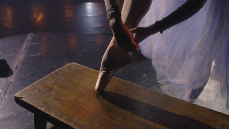 Hermosa-Bailarina-De-Ballet-Vestida-Se-Pone-Y-Ata-Zapatos-De-Punta-Para-El-Ensayo-De-Coreografía.-Prácticas-De-Bailarina-En-El-Escenario-Del-Teatro-Iluminado-Por-Focos.-Espectáculo-De-Danza-De-Ballet-Clásico.