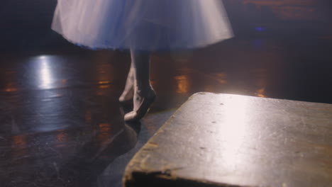 Ballerina-puts-on-pointe-shoes-for-choreography-rehearsal.-Ballet-female-dancer-practices-before-performance-and-dances-on-tiptoe-on-theater-stage-illuminated-by-spotlights.-Classical-ballet-dance.