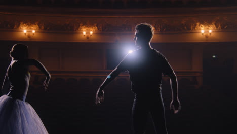 Man-in-training-suit-lifts-ballerina-on-his-arms-and-spins-around-himself-on-theatre-stage.-Pair-of-classical-ballet-dancers-on-rehearsal-before-performance.-Illuminated-theatrical-hall.-Slow-motion.