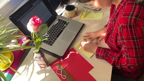 Frau-Schreibt-Auf-Einem-Papier-Aus-Einem-Notizbuch-Auf-Dem-Schreibtisch,-Während-Sie-Zu-Hause-Im-Iran-Arbeitet.-Teheraner-Iraner-Lernen-Persische-Sprache-In-Pandemie-Online-Kursen,-Klasse,-Heimdekoration,-Design,-Rotes-Kleid,-Rosa-Blume