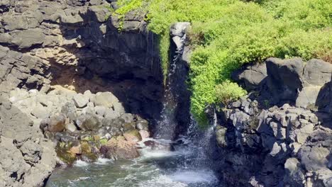 Cinematic-close-up-booming-down-shot-of-a-waterfall-flowing-into-a-swirling-tidepool-among-ancient-lava-rocks-near-Queen's-Bath-on-the-coast-of-Kaua'i,-Hawai'i