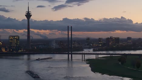 Barges-sail-on-flooded-Rhine-river-through-Dusseldorf-at-dusk,-Telephoto-drone-shot