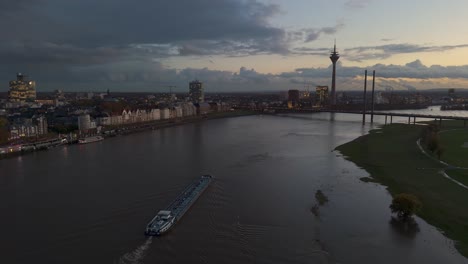 Barge-sails-up-flooded-Rhine-river-at-dusk,-Dusseldorf,-drone-tracking-shot