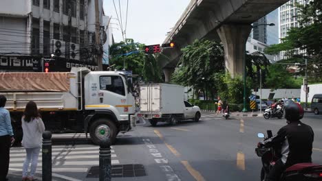 Eine-Motorradlieferung-Fährt-Vorbei,-Ein-Motortaxi-Mit-Einer-Frau,-Die-Ihr-Mobiltelefon-Benutzt,-Während-Andere-Vor-Dem-Grünen-Ampelschild-Darauf-Warten,-Dass-Alle-Eine-Belebte-Straße-In-Sukhumvit,-Bangkok,-Thailand,-überqueren