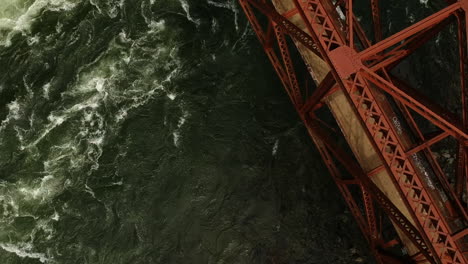 Ariel-drone-shot-of-raging-river-crossing-under-red-steel-bridge